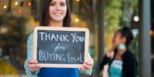 young business owner holding a thank you for buying local sign in front of her store with shopper in the background